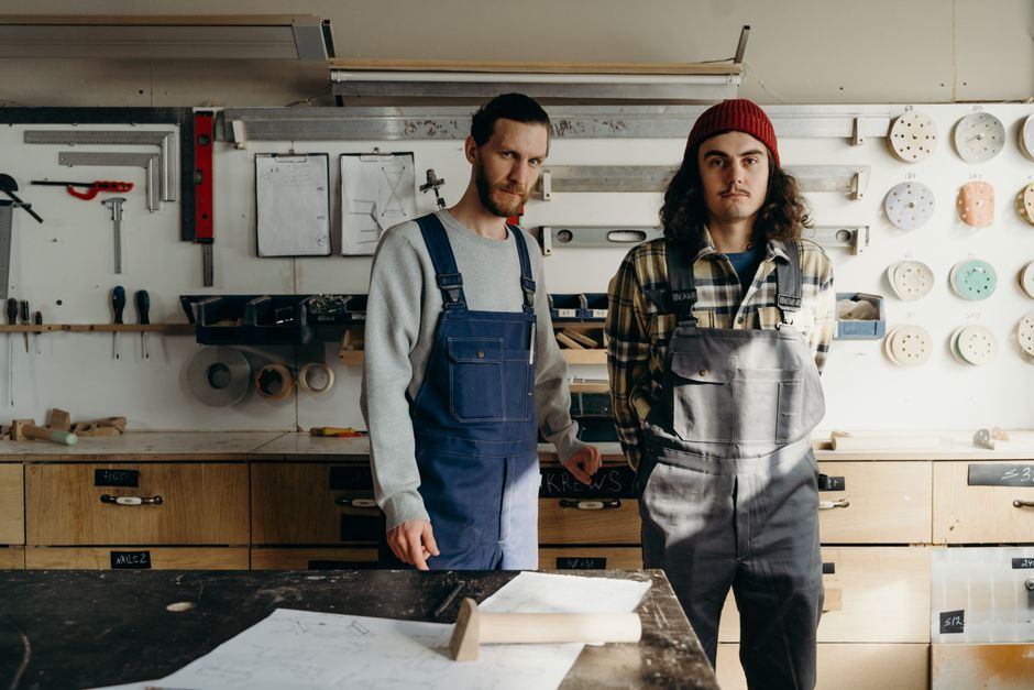 Two carpenters in overalls stand in a well-equipped workshop, surrounded by tools.